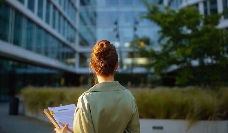 A woman standing outside a modern office building holding a clipboard with an agreement.