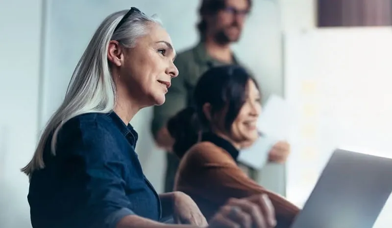 A woman with gray hair working in a collaborative setting with colleagues in the background.