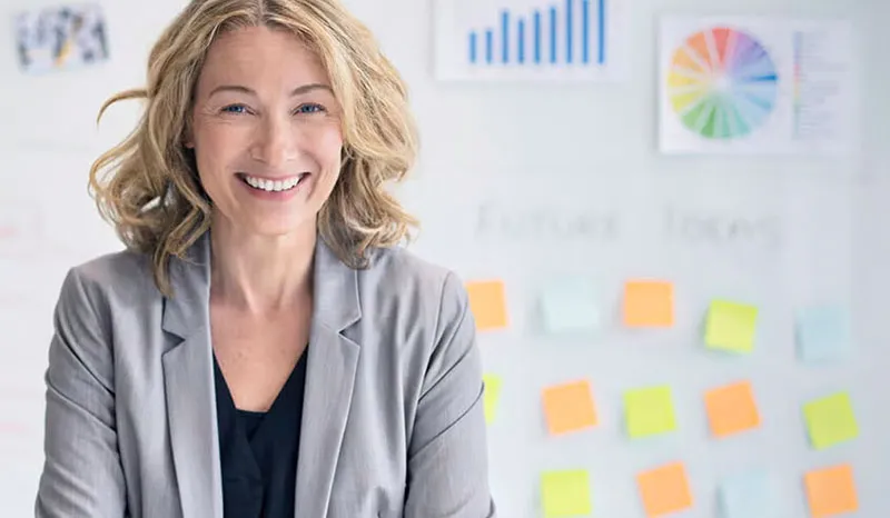 A professional woman smiling confidently while standing in an office, with colorful sticky notes in the background.